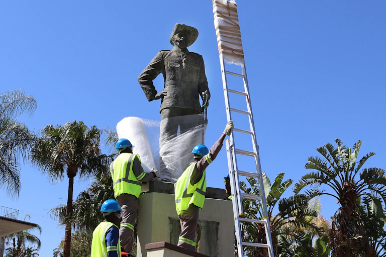 Men in yellow jackets and helmets climb the plinth of a statue of a man with a hat and an arm resting on his hip. They are wrapping the statue in bubble wrap.