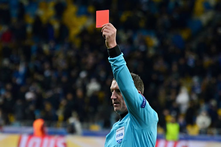 A football referee in a light blue jersey holds up a red card during a match