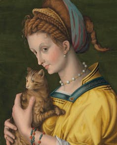 A painting of a young woman in a yellow dress, her hair wrapped in fabric and a pearl choker round her neck, holding a tabby kitten to her chest in a pose of affection.