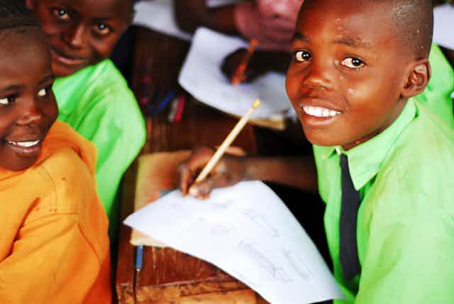 A boy smiling at the camera as he holds a pencil above a piece of paper in a classroom as two of his classmates in the shot smile at him.