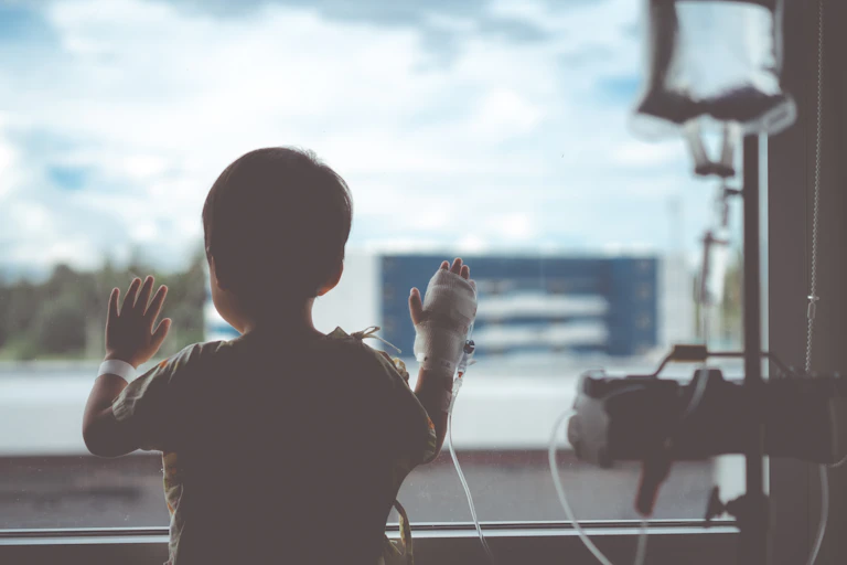 Un niño de espaldas mira por una ventana. Tiene una mano vendada y enchufada a un aparato médico.