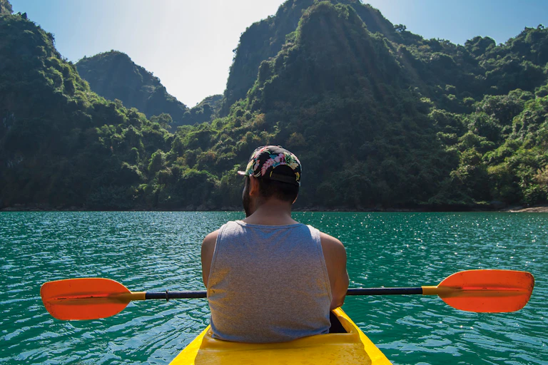A man paddling a yellow kayak in bright blue sea surrounded by picturesque mountains