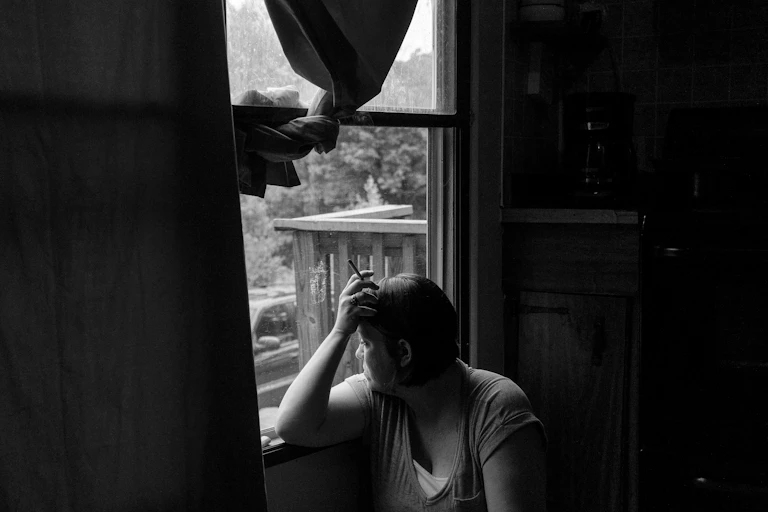 Black and white photo of a woman looking over her shoulder and out a window while holding a cigarette in her hand