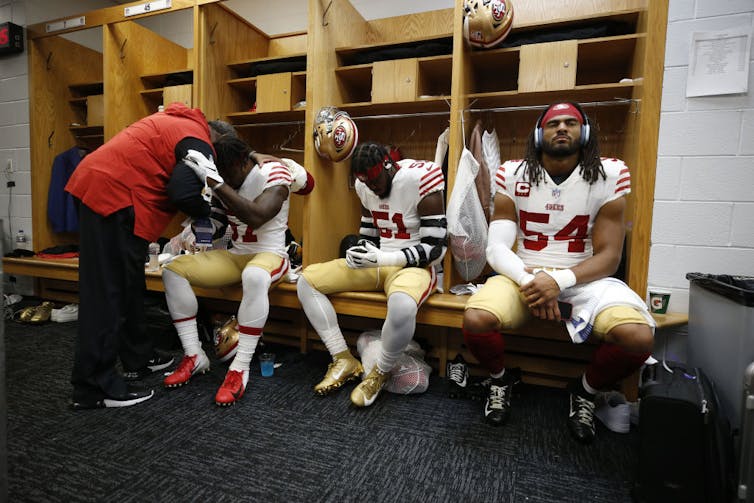 A man in an orange vest bends over as he prays with a man in a football uniform, seated next two other players in a locker room.