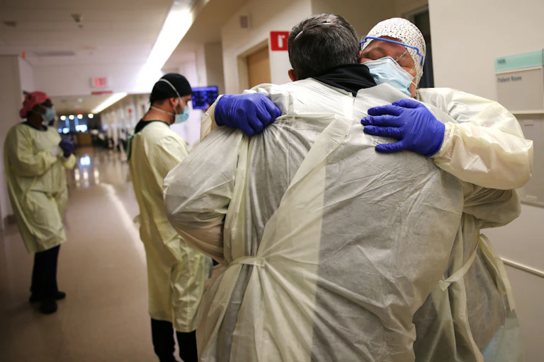 Two people in hospital garb and masks embrace inside a hallway.