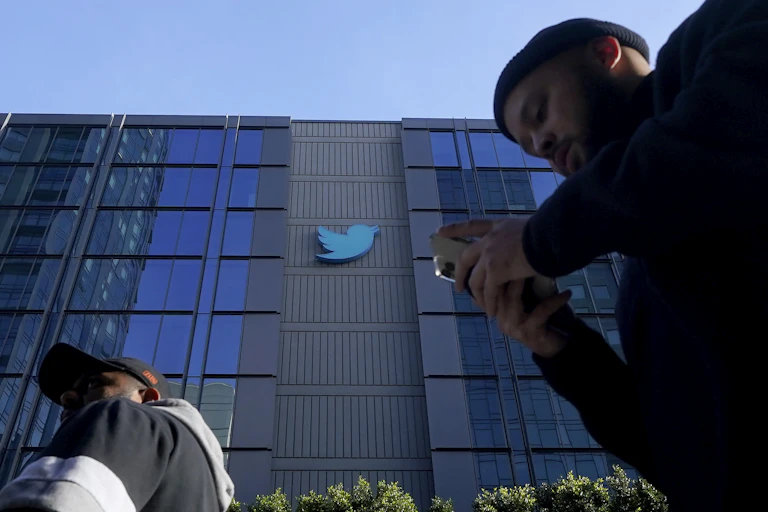 A person seen walking in front of the Twitter building holding a phone.
