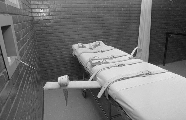 A black and white photo shows a white gurney with straps in a bricked room.