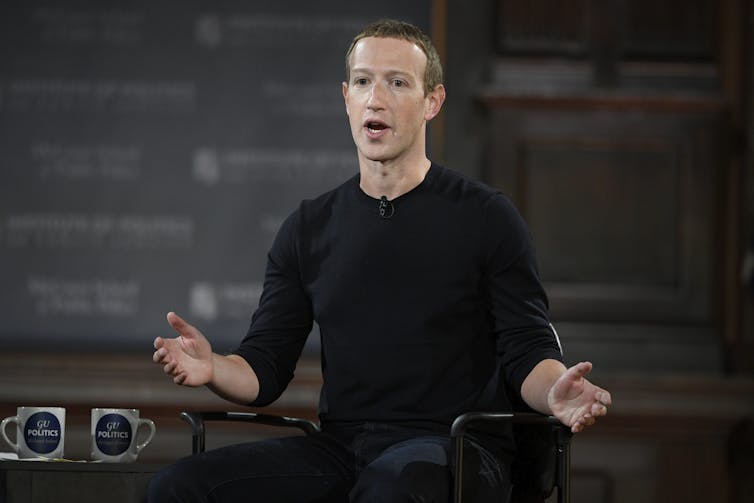 A white man in a black long-sleeved shirt gestures while speaking