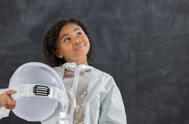 A young Black girl in a white space suit holds a space helmet as she looks skyward.