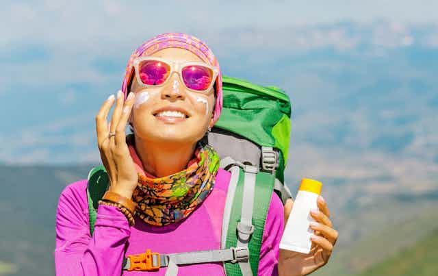 A woman in bright colourful hiking clothes and pink sunnies applying sunscreen to her face