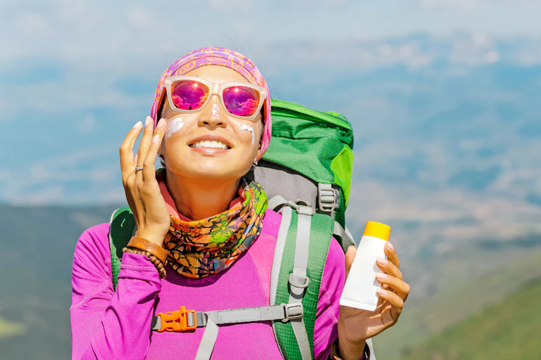 A woman in bright colourful hiking clothes and pink sunnies applying sunscreen to her face
