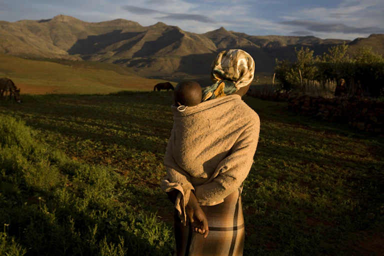 A woman with a baby on her back looking into a scenic landscape with hills and farmland.