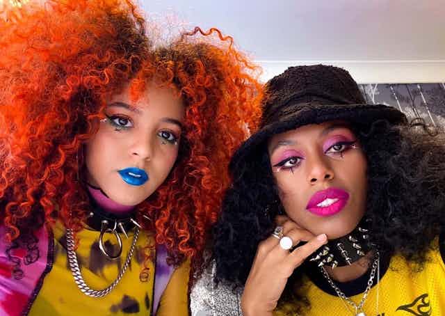 On the left, Georgia has voluminous orange curly hair, blue lipstick and a spiky dog collar. On the right, Amy wears a black bucket hat and pink lipstick. 