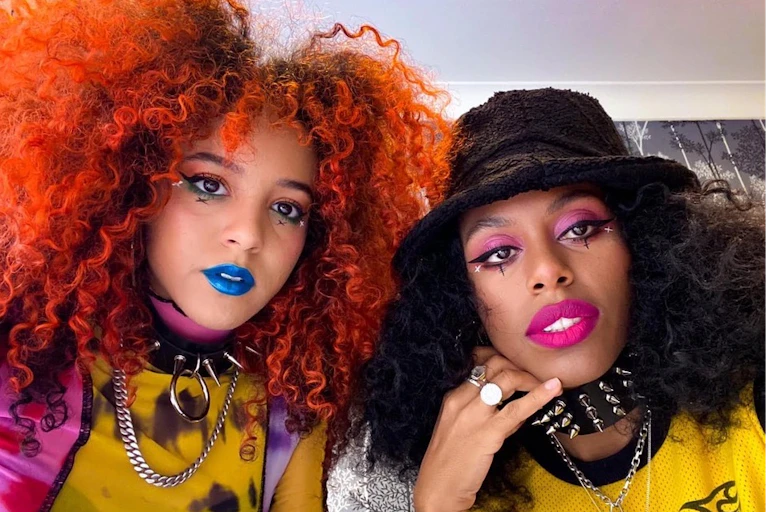 On the left, Georgia has voluminous orange curly hair, blue lipstick and a spiky dog collar. On the right, Amy wears a black bucket hat and pink lipstick.
