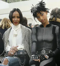 Jada and Willow sit side by side at a fashion show, Jada in a white shirt and Willow in a futuristic grey bodysuit.
