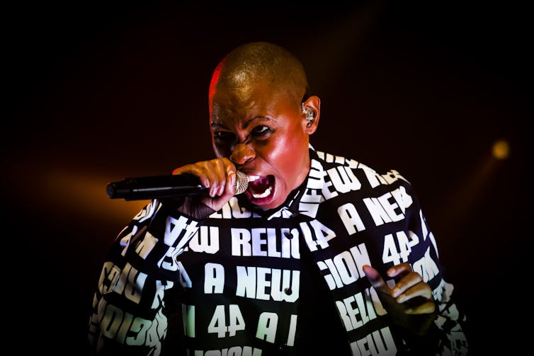 With a shaved head and bold black and white jacket, Skin sings into a microphone.