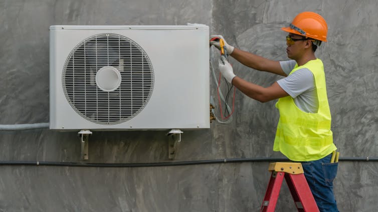 An engineer in high-visibility jacket installs an air conditioning unit to the exterior of a building.