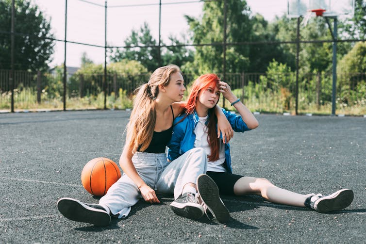 Two girls in sports clothes and with a basketball are chatting, sitting on the playground.