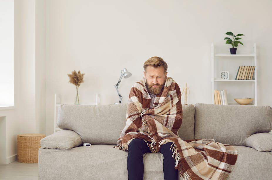 A man with a beard sitting on the sofa with a blanket wrapped around him.