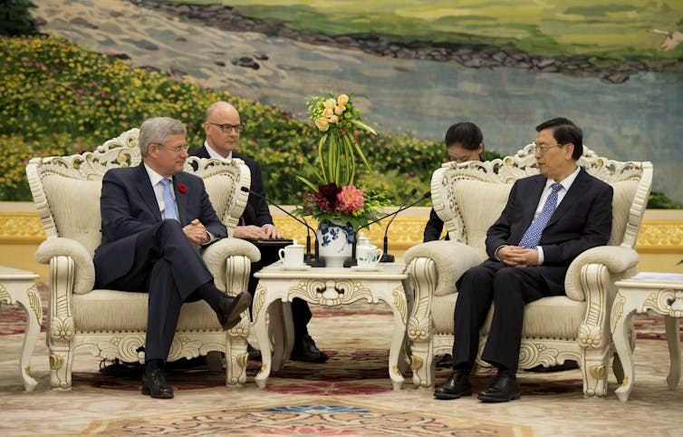 A man in glasses sits talking to an Asian man in an ornate setting with a large painted mural behind them.