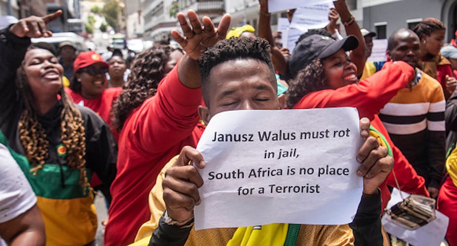 Men and women raising their hands in protest, with one man holding a sign that says 'Janusz Walus must rot in jail, South Africa is no place for a terrorist'.