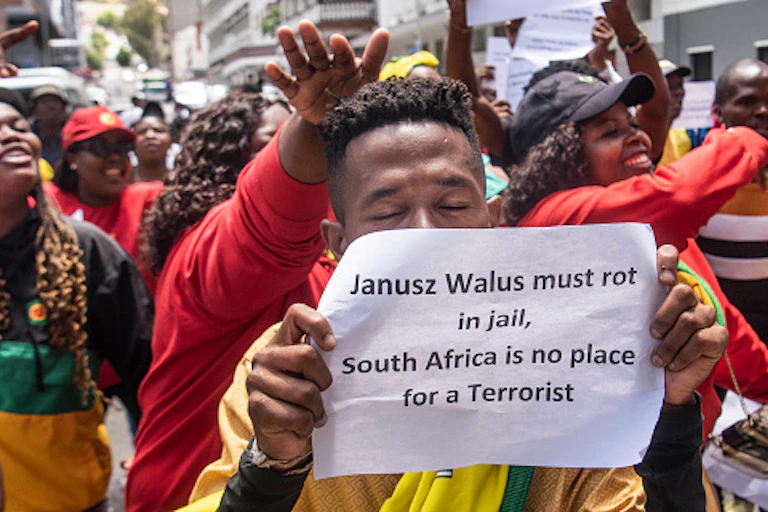 Men and women raising their hands in protest, with one man holding a sign that says 'Janusz Walus must rot in jail, South Africa is no place for a terrorist'.