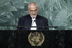 Un homme chauve parle dans un microphone sur un podium portant l’insigne de l’ONU