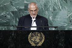A bald man with glasses speaks into a microphone at a podium with the UN insignia.