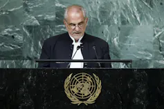 A bald man with glasses speaks into a microphone at a podium with the UN insignia.