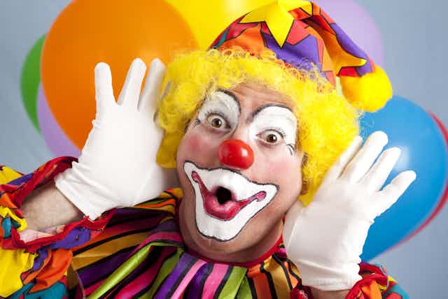 A male clown with a surprised expression and balloons in the background
