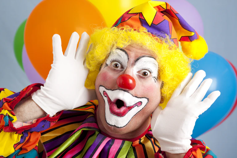 A male clown with a surprised expression and balloons in the background