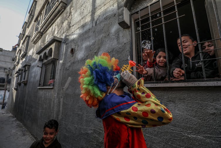A clown in a face mask blows bubbles while children grin behind a barred window