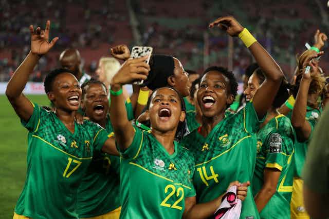 A group of women in South African football outfits shout and smile as a woman takes a selfie of them on a football field.