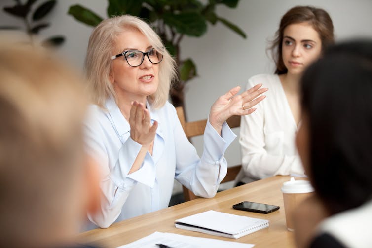 A woman wearing glasses and a white shirt speaking to other people around a table.