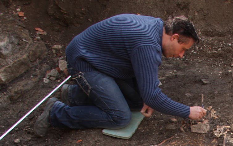 a man kneeling on pad on dirt works on something buried in the ground
