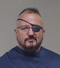 A balding, bearded man wearing glasses and an eye patch.