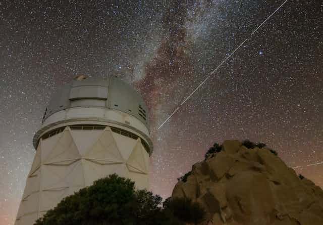A long exposure of the night sky, showing a white telescope tower with milky way in the background, and a bright white streak across the sky