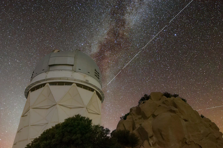 A long exposure of the night sky, showing a white telescope tower with milky way in the background, and a bright white streak across the sky