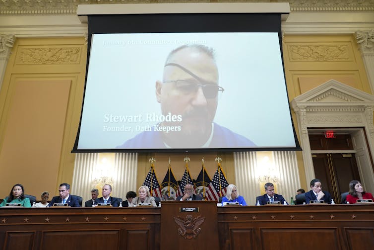 A man with an eyepatch shown on a large screen behind a group of people seated at a long desk in a public hall.