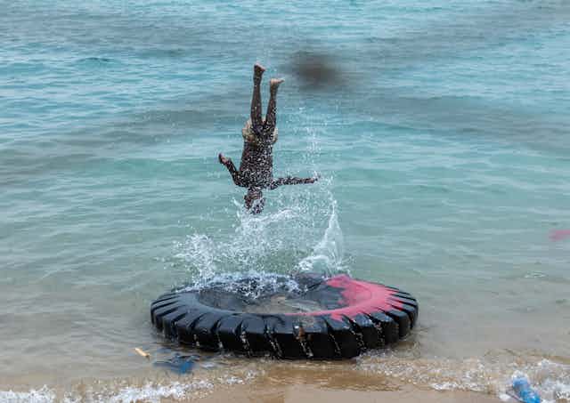 A boy turns a somersault above a large tyre partially submerged in blue-green seawater