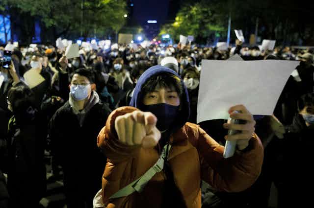 A protester in a balaclava holding a blank sheet of paper points at the camera.