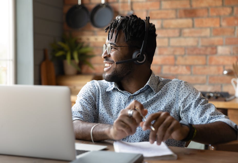 Man wearing headphones sitting at table with laptop, looking to side, smiling.