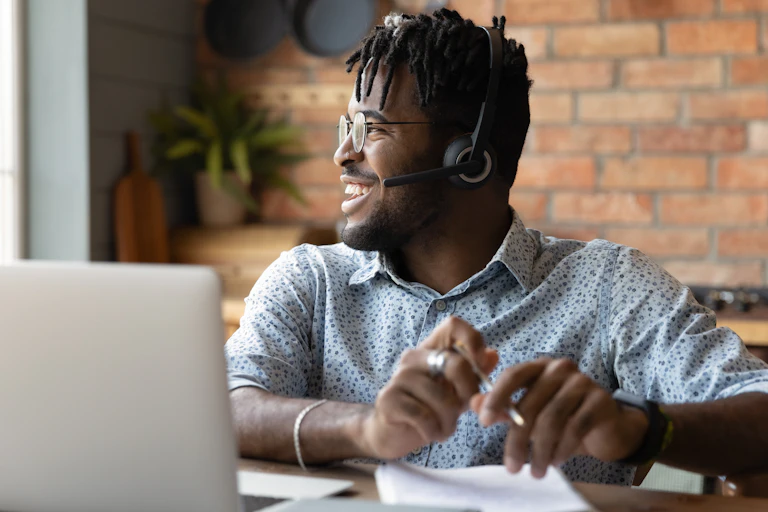 Man wearing headphones sitting at table with laptop, looking to side, smiling.