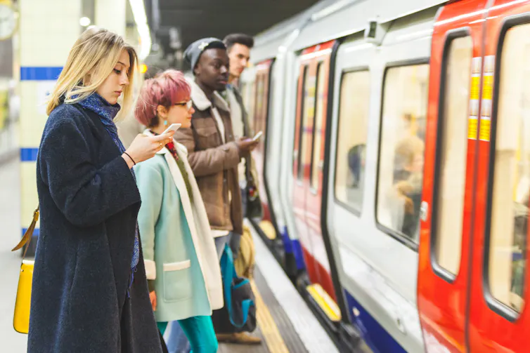 Two men and two women, standing on line, waiting to board a train, checking phone.