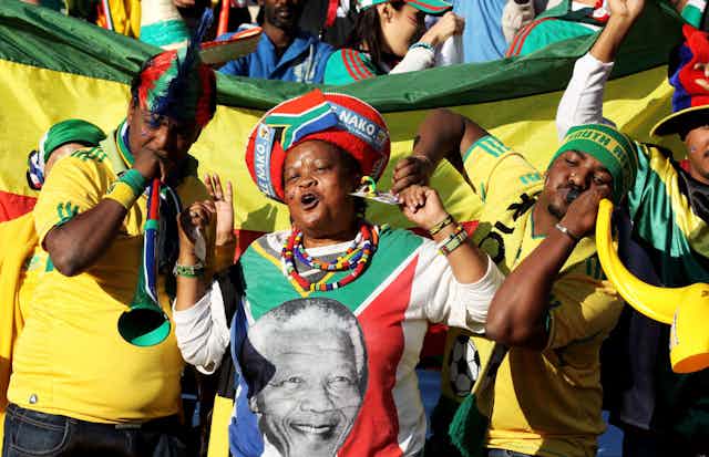 Three people stand in front of a flag among a crowd. They are brightly dressed in clothes that bear the South African flag and its colours. Two men blow on vuvuzelas and a woman has traditional headgear and an image of Nelson Mandela on her T-shirt, full of joy.