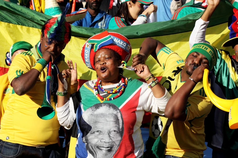 Three people stand in front of a flag among a crowd. They are brightly dressed in clothes that bear the South African flag and its colours. Two men blow on vuvuzelas and a woman has traditional headgear and an image of Nelson Mandela on her T-shirt, full of joy.