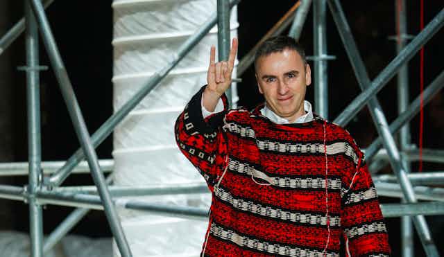 Raf Simons makes the rock symbol with his hand aloft, wearing a striped loose yarn baggy red jumper. 