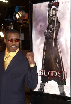 Man with red sunglasses pumps his first in front of a movie poster.