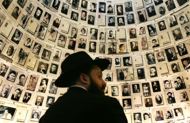 A white man wears a dark hat and black jacket and looks over his shoulder, as he faces a large wall of black and white, old looking photographs of people.