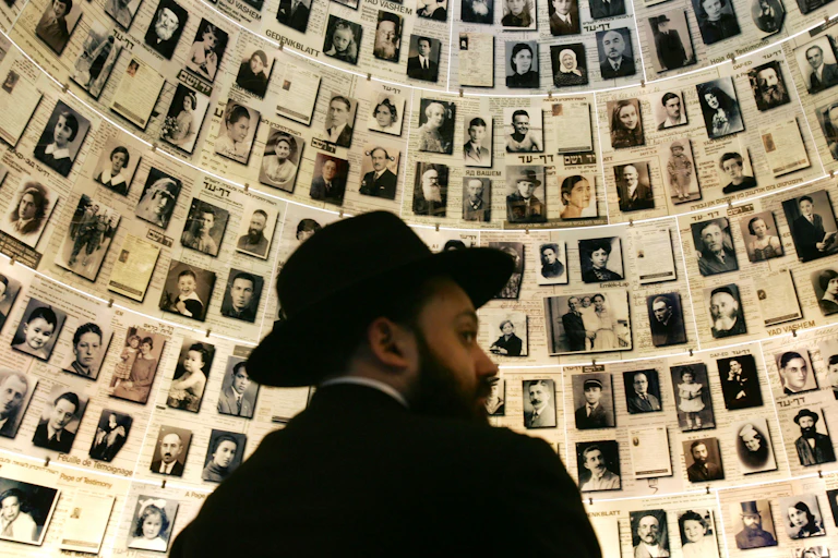 A white man wears a dark hat and black jacket and looks over his shoulder, as he faces a large wall of black and white, old looking photographs of people.